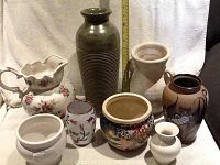 Lot of nine porcelain and ceramic vases, pots, and water jugs in various shapes and painted designs, placed on white fabric with measuring tape showing height of tallest vase.