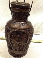 Photo showing front side of copper urn with carved figure holding a shield and spear.