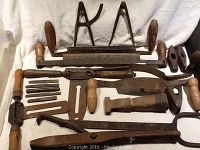 Full view of the 19 antique woodworking tools laid out on light fabric background showing wooden-handled hand planes, marking gauges, knives, and other metal tools with rust patina.