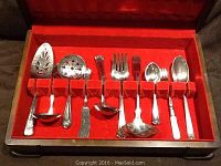Ten pieces of unmatched silverplate serving utensils displayed inside a wooden box with red felt lining.