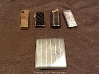 Four vintage butane lighters in different designs placed above the silver cigarette case on a cloth surface.