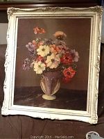 Framed floral print showing bouquet of diverse flowers in a classical vase in an ornate cream frame with wear.