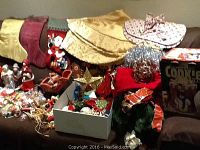 Photo showing a variety of Christmas stockings, a gold tree skirt, a snowman cookie jar, and a box with numerous ornaments.
