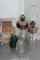 Photo showing two glass oil lamps with metal burners, two glass hurricane lamp shades, boxed pair of cut glass oil lamps, and two glass insulators in front.