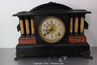 Front view of black Ingraham Co mantle clock with decorative faux marble columns and brass feet. Clock face is circular with black numbers and gold center.