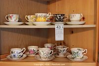 Twelve assorted tea cups and saucers displayed on two wooden shelves, various floral and colorful patterns visible.