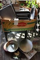 Box containing multiple metal baking pans in various shapes and sizes, with some strainers and a wooden rolling pin visible in front.