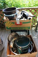 Boxes filled with assorted cookware including sauce pans, fry pans, and roasting pans stacked on and beside a small patio table outside.