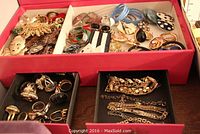 Open pink jewelry box showing assorted costume jewelry pieces including earrings, pins, rings, and bracelets laid inside compartments and trays.