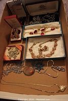 Photo showing multiple jewelry items including boxed pins, necklaces, bracelets, and cuff links in a cardboard box