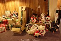 Wide view of assorted Christmas decorations including plush toys, boxes of lights, and figurines arranged on the floor and against wall