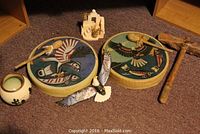 Two round hand drums with native bird designs lie on the floor along with a wood-carved eagle figurine, small pottery, miniature adobe-style structure, wooden drumstick, and rustic wood hatchet, all reflective of southwest native decor.