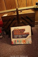 Closed wooden sewing cabinet with carrying handle and folding compartments shown with Reader's Digest Complete Guide to Needlework book placed in front.