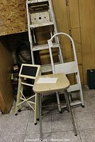 Overall view of two aluminum ladders, two step stools, and a padded metal stool, placed in a room with brown wall and tiled floor.