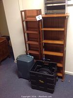 Three wooden DVD racks arranged side by side along the wall, with a grey vertical paper shredder and a black rolling cart with folding handles placed in front of them on a carpeted floor.