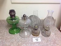 Four vintage hurricane lamps on a countertop. The tall lamp on the left is green glass missing the glass chimney (globe). The other three lamps have clear glass bases with metal burners and intact glass chimneys. One chimney has frosted etched sunburst design.