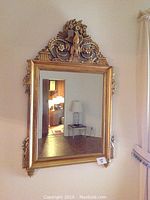 Front view of large rectangular wall mirror with an ornate gold-toned wooden frame. Mirror reflects part of room and furniture.