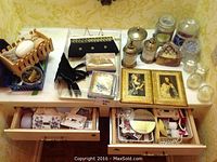 View of ladies evening bags, framed portraits, metal containers, glass jars, and various decor and vanity items spread on a vanity top with open drawers containing assorted small items.