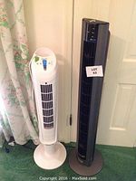Photo showing two tower fans standing side by side on a green carpet. Left fan is white, smaller; right fan is black, taller with remote and control panel on top.