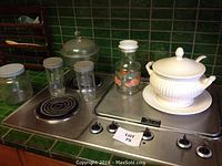 White ceramic soup tureen on metal stove surface with several glass jars and containers