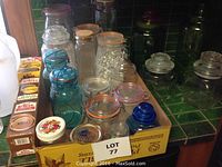 Variety of canning jars and lids, including blue bail wire jars and clear glass jars with metal lids. Displayed collectively in a box showing assortment of jar types and lids.