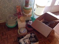 Various plastic bowls, containers, kitchen utensils, and a cardboard box with miscellaneous items grouped on the floor against a wallpapered wall.