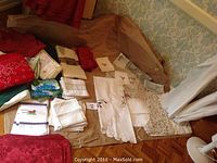 Overview of assorted linens including white cut work tablecloths, napkins, and placemats laid out on a brown sheet on the floor beneath window with white curtains.