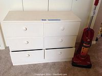White chest of drawers with six drawers and a red Dirt Devil vacuum cleaner standing on carpet.