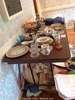 Photo of a brown portable folding table loaded with various dishes and glassware items, with tiled floor visible.
