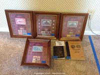 Photo showing four wooden-framed coin and stamp art pieces, each with a maroon background and multiple coins and stamps, plus two coin informational booklets on the floor.