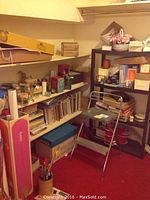 Photo of corner with metal step stool, black shelf unit with gift wrap items, and white wall shelves with books and miscellaneous decor.