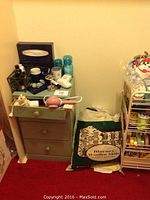 Small wood chest with three drawers, various small bottles and cups on top, bag of fabric or yarn to side, and a metal gift wrap cart with ribbons and wrapping supplies.