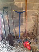 Photo showing a group of garden hand tools including rakes, shovel, pitchfork, and cultivator alongside a red gas-powered weedeater on the ground near a wall.