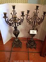 Front view of two vintage metal candelabras, showing one with complete arms and one with a missing arm, placed on tiled floor against white backdrop.