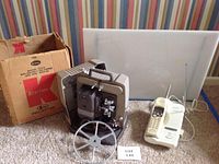 Photo shows a vintage Keystone 8mm projector with large reel and original box, a white light box standing vertically behind it, and a cream-colored Panasonic cordless phone with extendable antenna placed on a carpeted floor.