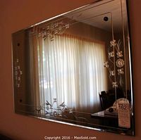 Full view of the rectangular mirror mounted on a wall reflecting a curtained window, showing the etched cornflower floral pattern at the corners and edges.