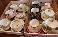 Twelve vintage tea cups and matching saucers arranged in two cardboard boxes, showing various floral and gold-trimmed designs.