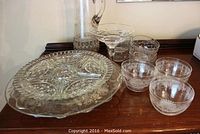 Pressed glass pitcher, silver overlay footed cake plate, cornflower platter, etched patterned fruit bowls shown together on wood surface.