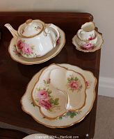 Close-up of teapot and small creamer and sugar bowl showing design details of the pink rose pattern and gold edging on white china.