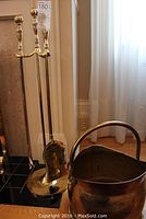 Full view of brass bucket and three brass fireplace tools (poker, shovel, brush) standing upright with handles topped with spherical finials.
