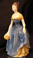 Front view of Royal Doulton porcelain figurine of a woman holding yellow flowers, dressed in a blue gown with yellow shawl and underskirt.