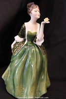 Frontal view of the Royal Doulton figurine showing the woman posed with her right hand raised holding a small flower, wearing a green dress with gold polka dots and floral designs at the hem.