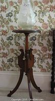 Mahogany plant stand with three splay legs and carved center column, holding vase, floral wallpaper background.