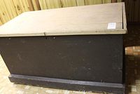 Side view of a painted wooden blanket chest showing dark brown body and light colored hinged lid with visible wear and scratches.