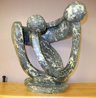 Full view of abstract Opalstone sculpture with two stylized figures in a curved embrace, displayed on a wooden surface against a plain background.