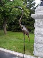 Front-on view of full-height heron sculpture standing on thin steel legs against stone pillar backdrop.