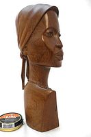 Side profile view of hand carved wooden bust showing woman with a tied headscarf and necklace, polished finish revealing Azora wood grain. Kiwi shoe polish tin for scale.