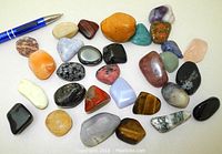 Image showing a collection of 30 polished semi-precious stones in various colors and shapes laid out on a white surface, with a pen for scale.