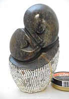 Front view of 'Lovers' serpentine stone sculpture showing two embracing abstract figures on textured base next to a small tin for size reference.