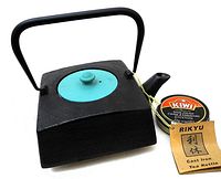 Front angled view of a square black cast iron tea pot with a turquoise round lid and handle. Tag with brand Rikyu and label noting it's a cast iron tea kettle, along with a Kiwi shoe polish tin for size reference.